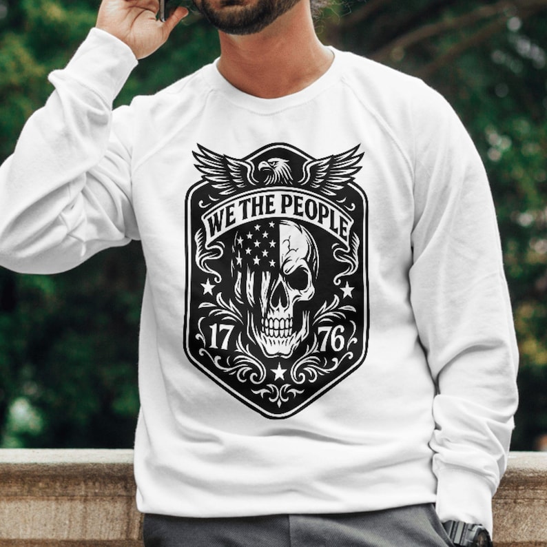 Puede incluir: Sudadera blanca con un gr&aacute;fico en blanco y negro. El gr&aacute;fico incluye una calavera con un dise&ntilde;o de la bandera estadounidense, el texto "WE THE PEOPLE", el a&ntilde;o "1776" y un emblema de &aacute;guila. La sudadera tiene mangas largas.