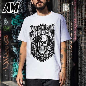 Puede incluir: Camiseta blanca con un dise&ntilde;o gr&aacute;fico en blanco y negro. El dise&ntilde;o incluye una calavera con un motivo de la bandera estadounidense, el texto "WE THE PEOPLE" y los n&uacute;meros "17" y "76". La camiseta tiene mangas cortas y cuello redondo.