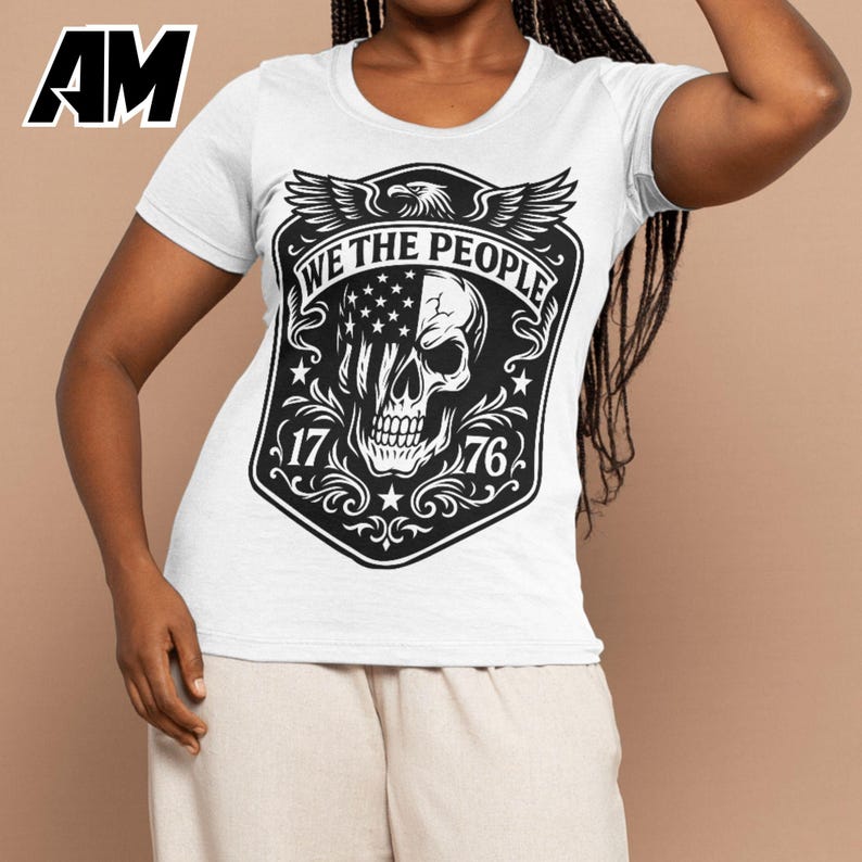 Puede incluir: Camiseta blanca con un dise&ntilde;o gr&aacute;fico en blanco y negro. El dise&ntilde;o incluye una calavera con la bandera estadounidense, las palabras "WE THE PEOPLE", los n&uacute;meros "17" y "76", y un &aacute;guila. La camiseta tiene mangas cortas.
