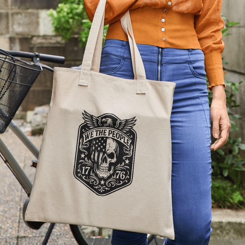 Puede incluir: Bolso tote de lona beige con un dise&ntilde;o gr&aacute;fico en blanco y negro. El dise&ntilde;o incluye una calavera, un &aacute;guila con las alas extendidas y el texto "WE THE PEOPLE" junto con los n&uacute;meros "17" y "76". El bolso tiene dos asas.