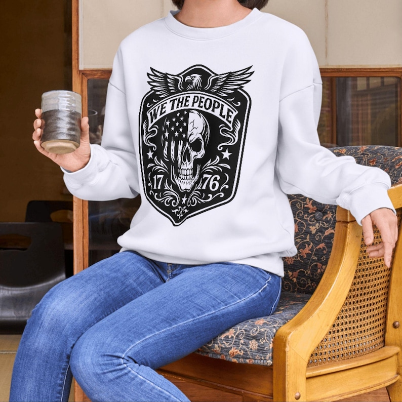 Puede incluir: Sudadera blanca con un dise&ntilde;o gr&aacute;fico en blanco y negro. El gr&aacute;fico incluye una calavera, un dise&ntilde;o de bandera estadounidense, las palabras "WE THE PEOPLE", los n&uacute;meros "1776" y un &aacute;guila. La persona lleva vaqueros azules y sostiene un vaso.