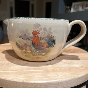 May include: A ceramic pitcher with a rooster and chicks design. The pitcher is beige with green trim and features a rooster with a red comb and black tail feathers. The rooster is surrounded by chicks and a green bush.