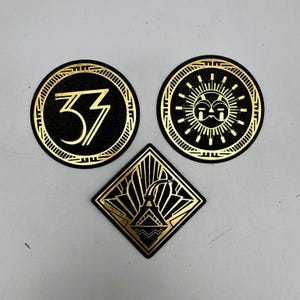 May include: Three black and gold patches. Two are circular, one with the number 33 and the other with a sun face design. The third patch is diamond-shaped with an art deco design.