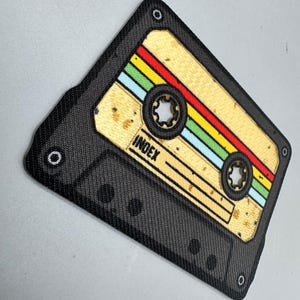 May include: A black cassette tape patch with a tan center and colorful stripes of red, yellow, green, and blue. The word "INDEX" is printed on the tape. The patch has a textured appearance and is edged with black.