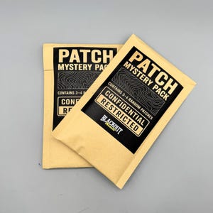 May include: Two tan paper envelopes labeled "PATCH MYSTERY PACK" with black text and a topographic design. The envelopes also state "CONTAINS 3-4 RANDOM PATCHES" and "CONFIDENTIAL RESTRICTED". The brand name "BLACKOUT threads" is visible.