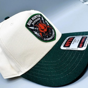 May include: A white and dark green baseball cap with a patch that reads "Bug Hunter Division" and a red and black graphic. The cap has a curved brim and a "Stylish Look" sticker.