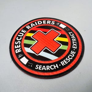 May include: Circular patch with the words "RESCUE RAIDERS" and "SEARCH RESCUE EXTRACT" in white text on a black background. A red "X" is in the center, surrounded by colorful stripes. The patch has a red border.
