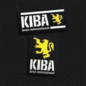 May include: Two rectangular black embroidered patches featuring the white text KIBA and the words Arms international in smaller white font. A yellow lion graphic holding a firearm is positioned next to the text on both patches.