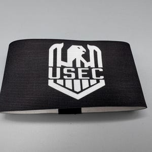 May include: Black armband with the white text "USEC" and a stylized eagle emblem. The armband is made of a ribbed fabric and has a simple, clean design. The text and emblem are centered on the band.