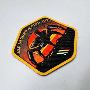 May include: Embroidered patch with a black climber silhouetted against a red and orange sunset. The patch has the text "ARC AROUND & FIND OUT" in white, with a yellow border and a small striped design.