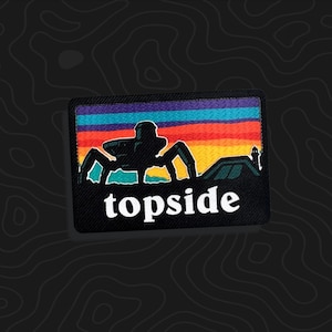 May include: A rectangular embroidered patch featuring a multi-colored sunset background with horizontal stripes of purple, blue, orange, and yellow. A dark silhouette of a multi-legged mechanical creature and a building appear above the word topside.