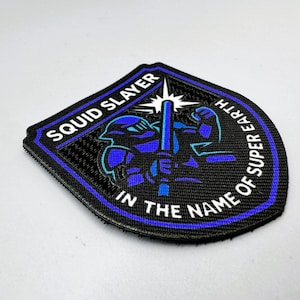 May include: Black and blue embroidered patch with the text "SQUID SLAYER" and "IN THE NAME OF SUPER EARTH". The patch features a blue warrior figure and a starburst design.