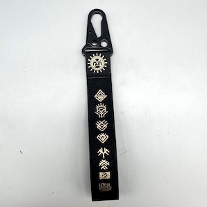 May include: Black keychain strap with gold-colored symbols and a black metal clasp. The strap features a sun design and various geometric and stylized symbols. The keychain is designed for attaching keys or other small items.