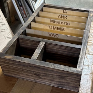 May include: A stained wooden organizer with multiple compartments. The organizer has vertical dividers and labeled sections with text including "IA", "ANK", "Missorts", "UMMB", and "VAC". The wood has a dark, weathered finish.