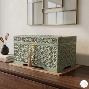 Op de afbeelding: Een saliegroene decoratieve kist met een geometrisch patroon, sleutel en kwast. De kist staat op een houten oppervlak, naast een kleine vaas met decoratieve stelen en een stapel boeken. Een goudomrandde wanddecoratie is zichtbaar op de achtergrond.