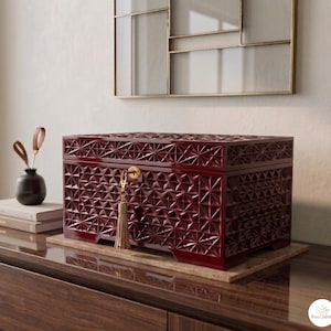 Puede incluir: Una caja de almacenamiento rectangular de color rojo oscuro con acabado brillante y un patrón geométrico en relieve en forma de estrella en todos los lados visibles. La caja cuenta con una cerradura dorada central con una borla beige.