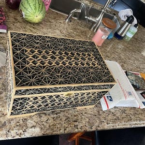 May include: A decorative wooden box with a geometric black and beige pattern. The box has a hinged lid and a gold-coloured clasp. The design features repeating star and diamond shapes, creating a textured appearance.