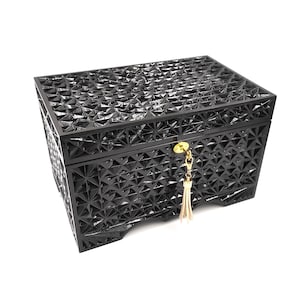 Puede incluir: Caja decorativa negra con un patrón geométrico de estrella. La caja tiene una tapa con bisagras y una llave dorada con una borla. La caja está hecha de un material oscuro y brillante.