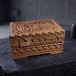 May include: A wooden box with detailed carvings and a combination lock. The rectangular box features a floral design on the lid and geometric patterns on the sides. The box is made of light brown wood and rests on a dark stone surface.