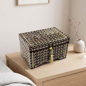 May include: A rectangular black storage box featuring an intricate gold-colored filigree pattern across its exterior surfaces. The box includes a central gold-colored lock mechanism with a hanging gold tassel and a hinged lid with a decorative design.