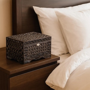 May include: A dark brown, intricately carved wooden box with a geometric pattern sits on a matching nightstand. The box has a small keyhole and is next to a bed with white bedding.