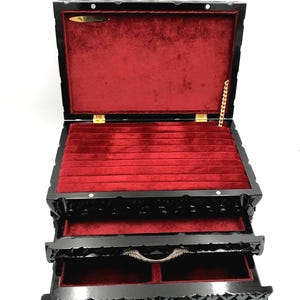 May include: Black jewelry box with a red velvet interior. The box has two pull-out drawers and a hinged lid. The exterior is intricately carved with a floral design. The box is open, revealing the interior compartments.