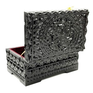 Può includere: Un portagioie nero aperto con intricati disegni floreali intagliati. L'interno è foderato in velluto rosso. La scatola è rettangolare con coperchio incernierato e chiusura color oro.