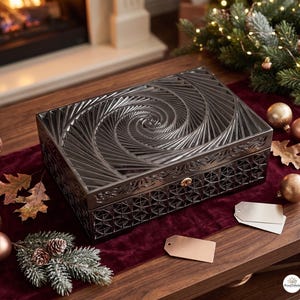 Könnte beinhalten: Eine rechteckige schwarze Aufbewahrungsbox mit einem tief geschnitzten Spiralmuster auf dem Deckel und einem geometrischen Gitterdesign an den Seiten. Die Box verfügt über einen kleinen, kreisförmigen goldfarbenen Verschluss an der Vorderseite.