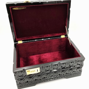 Può includere: Baule in legno nero aperto con interno in velluto rosso. Il baule ha un motivo decorativo intagliato all'esterno e un fermo color oro. La serratura a combinazione mostra il numero 1.