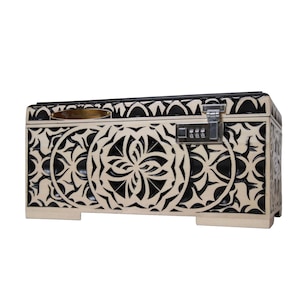 May include: A rectangular chest with a cream-colored exterior and intricate black cut-out designs. It features a combination lock and a gold-colored handle. The chest is elevated on small feet.