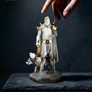 Peut inclure: Une statue de style marbre blanc représentant Hadès avec des accents dorés sur la couronne, les épaulettes, la ceinture et le trident. La figurine est accompagnée d'un chien à trois têtes enchaîné et porte le texte : for the modern deity. Our Hades statue is an authentic, collectible sculpture. Forged from myth, designed for your domain.