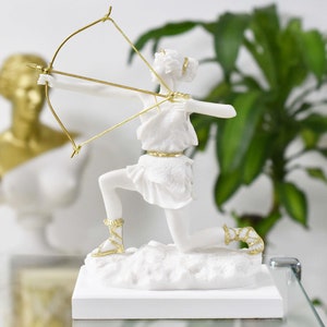 May include: White marble statue of a woman kneeling and holding a bow and arrow. The statue has gold accents on the bow, arrow, and clothing.