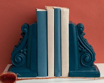 May include: A dark blue bookend set with a decorative design. Three books are displayed in the bookends, two are white and one is blue.