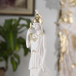 Può includere: Statua bianca di una figura femminile con un elmo dorato e che tiene una lancia. La statua è decorata con accenti dorati e ha uno sfondo bianco.