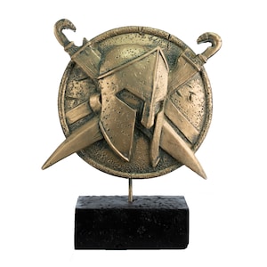 May include: A gold-colored metal sculpture of a Spartan helmet with two crossed swords on a round shield. The sculpture is mounted on a black base.