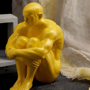 May include: A yellow, stylized sculpture of a person sitting with their knees pulled up to their chest. The sculpture is made of a smooth, glossy material.