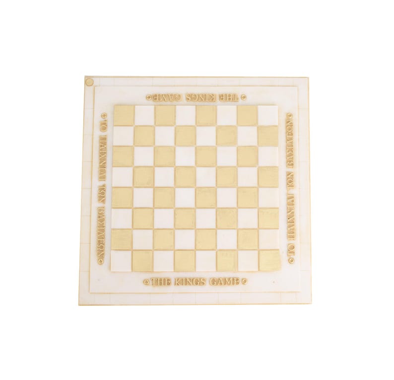 K&ouml;nnte beinhalten: Ein quadratisches Schachbrett mit zweifarbigem Schachmuster in Gold und Wei&szlig;. Das Brett tr&auml;gt den Text "THE KINGS GAME" und griechischen Text um den Rand. Das Brett besteht aus einem hellfarbigen Material.