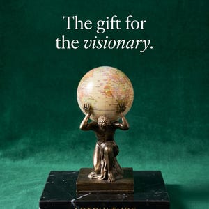 Op de afbeelding: Een bronskleurig beeld van een knielende figuur die een wereldbol op de schouders draagt, geplaatst op een rechthoekige voet van zwart marmer. De tekst ART CULTURE en The gift for the visionary is in witte letters boven het beeld zichtbaar.
