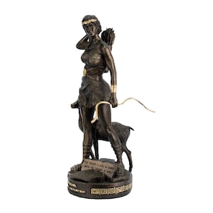 May include: A bronze statue of a woman holding a bow and arrow, standing next to a fawn. The statue is on a base with the inscription "The moon learns a smile when she learns a trick".