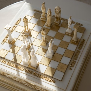 K&ouml;nnte beinhalten: Ein dekoratives Schachspiel mit gold-wei&szlig;en Feldern und detaillierten Figuren. Das Schachbrett tr&auml;gt goldfarbene Schriftz&uuml;ge: "THE KINGS GAME" und "&Tau;&Omicron; &Pi;&Alpha;&Iota;&Chi;&Nu;&Iota;&Delta;&Iota; &Tau;&Omega;&Nu; &Beta;&Alpha;&Sigma;&Iota;&Lambda;&Epsilon;&Omega;&Nu;". Die Figuren sind wei&szlig; und gold.