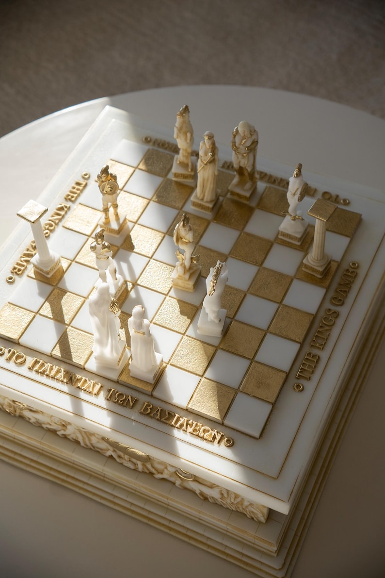 K&ouml;nnte beinhalten: Ein dekoratives Schachspiel mit gold-wei&szlig;en Feldern und detaillierten Figuren. Das Schachbrett tr&auml;gt goldfarbene Schriftz&uuml;ge: "THE KINGS GAME" und "&Tau;&Omicron; &Pi;&Alpha;&Iota;&Chi;&Nu;&Iota;&Delta;&Iota; &Tau;&Omega;&Nu; &Beta;&Alpha;&Sigma;&Iota;&Lambda;&Epsilon;&Omega;&Nu;". Die Figuren sind wei&szlig; und gold.