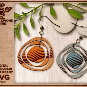 May include: A pair of laser-cut wooden earrings. One earring is a blue and brown geometric design, the other is orange and brown. The earrings are on a wooden background with green leaves.