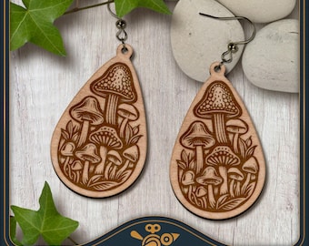 May include: A pair of teardrop-shaped wooden earrings featuring a laser-engraved design of various mushrooms and foliage. The earrings are suspended from silver-toned metal hooks and display a detailed, dark brown illustration on a light wood surface.