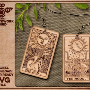May include: A pair of rectangular wooden earrings featuring laser-engraved tarot card designs. One earring depicts The Sun with a central face and a figure on a horse, while the other shows The Moon with a crescent face, two wolves, and a lobster.