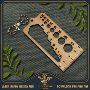 Puede incluir: Una herramienta de llavero de madera con una regla, tamaños de agujeros y un llavero. La herramienta tiene marcas de M2 a M12 y números del 0 al 19. El texto "LASER-READY DESIGN FILE" y "CLOCKWORK BIRD" son visibles.