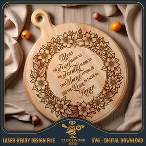 May include: A round wooden cutting board with a handle featuring a laser-engraved floral wreath surrounding the text: Bless the food before us, the family beside us, this home around us, and the love between us. Amen. Two small tomatoes are visible.