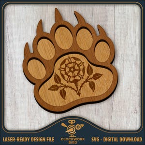 May include: A laser-cut wooden bear paw print featuring a central engraved Tudor rose design. The object consists of a dark brown outer frame with a lighter wood grain inlay for the paw pads and the detailed floral motif in the center.