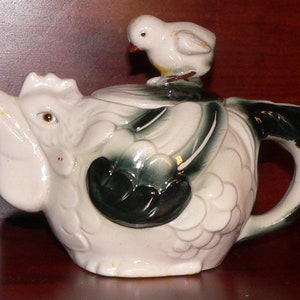 May include: White ceramic teapot shaped like a chicken with a small white bird perched on top. The chicken has green and black painted details on its wings and tail feathers.