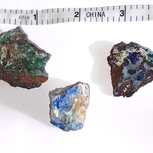 May include: Three raw mineral specimens with a ruler in the background. The minerals display a variety of colors, including green, blue, brown, and white. The ruler shows measurements in inches.
