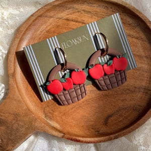 Pode incluir: Um par de brincos de argola com cestas trançadas castanhas cheias de maçãs vermelho-brilhante e pequenas folhas verdes. Os brincos estão presos a um cartão de apresentação verde com riscas brancas verticais e o texto FLOAKSUN.
